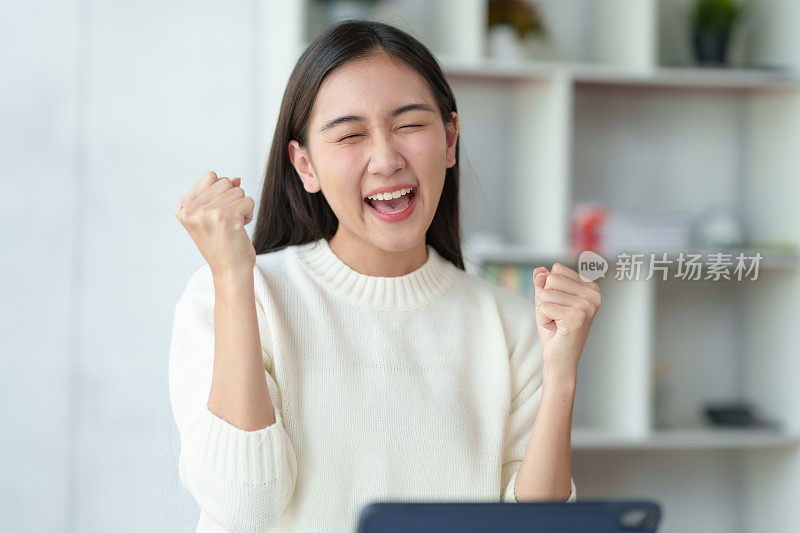 年轻的亚洲女性在笔记本电脑上工作，很高兴取得了满意的结果，并对完成的工作感到满意。