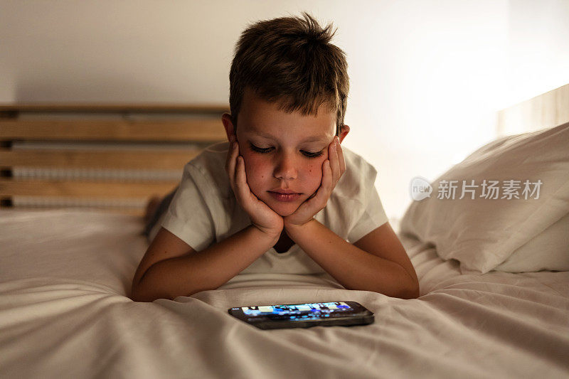 可爱的小男孩坐在黑暗中用手机看动画片。可爱的孩子晚上拿着智能手机躺在床上的特写。技术的概念。副本的空间。