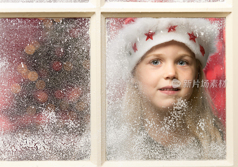 戴着圣诞帽透过白雪覆盖的窗户看圣诞节的女孩