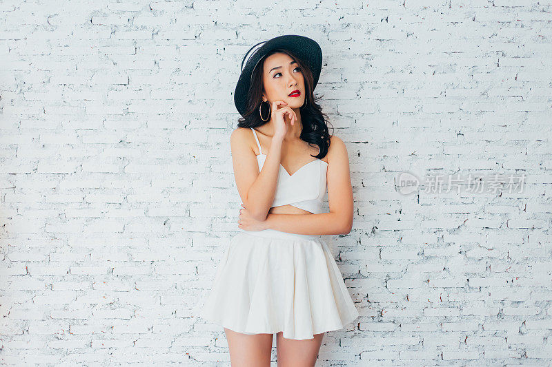 年轻女子想一些与时尚的夏季帽子白色墙壁背景与复制空间