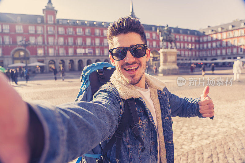 有吸引力的年轻白人旅游学生男子背包有乐趣快乐和兴奋的自拍与智能手机在广场市长，西班牙马德里。欧洲旅游、度假的概念。