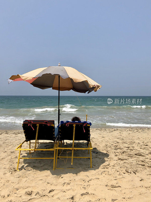 一对夫妇在印度南部喀拉拉邦的沙滩上享受夏日日光浴，金属黄色的太阳躺椅上有靠垫，热带海边和绿松石色的海景，海浪和潮汐，用遮阳伞遮挡炎热的太阳，防止晒伤