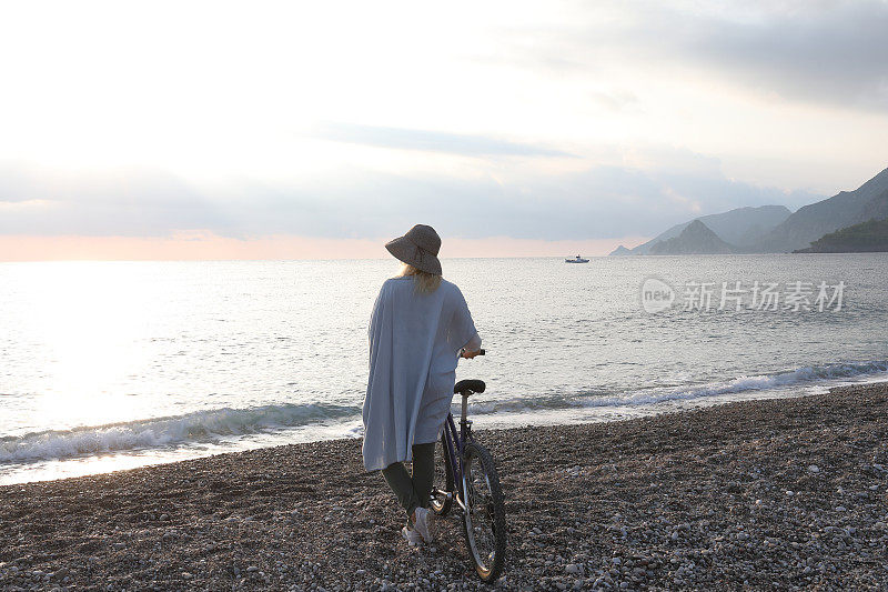 成熟的女人骑着自行车在日出的海滩上放松