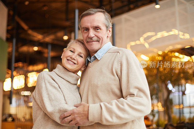 在充满灯光的现代餐厅里，一对幸福的老年夫妇穿着休闲毛衣互相拥抱