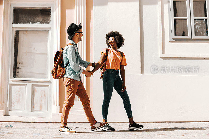 年轻可爱的微笑跨文化潮夫妇手牵着手走在街上，在阳光明媚的日子里玩得很开心。多样性的概念。