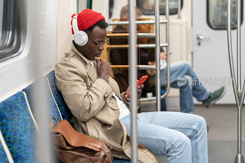 一名非裔美国乘客坐在地铁里，在公共交通工具上使用手机，戴着无线耳机听音乐。