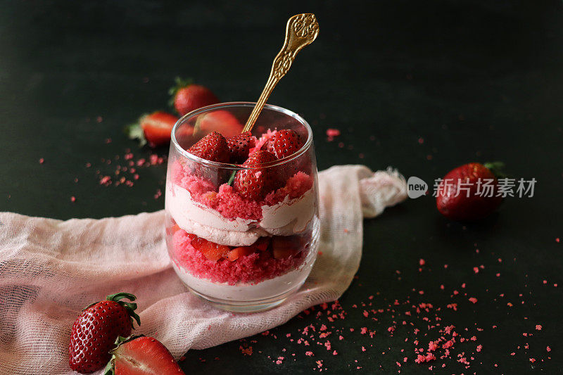 近摄图像玻璃包含自制的草莓慕斯甜点，上面覆盖着整个草莓，金叉，鲜奶油慕斯分层布丁和红色天鹅绒蛋糕屑，周围环绕着水果，粉红色的平纹布，黑色背景，聚焦前景