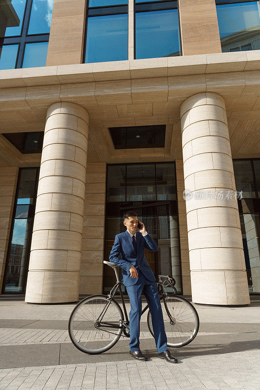 一位身穿蓝色西装的年轻亚洲商人斜靠在自行车上，靠在有柱子的现代商业中心的正面打电话