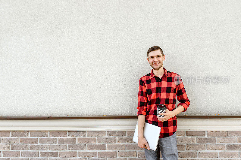 一个快乐的自由职业者站在一堵砖墙旁，手里拿着一杯咖啡和一台笔记本电脑