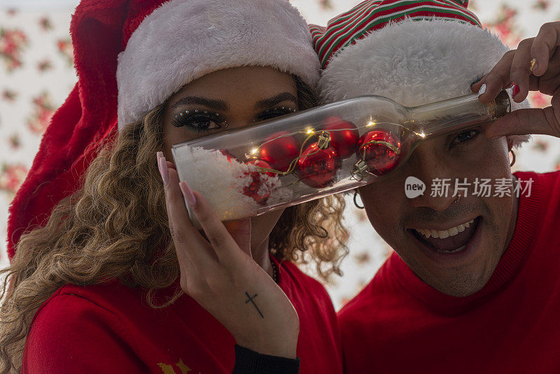 一对拉美裔黑人夫妇穿着红色的圣诞麻袋，戴着圣诞帽，背景是白色的铃铛，他们戴着圣诞帽站在一起，互相表达爱意
