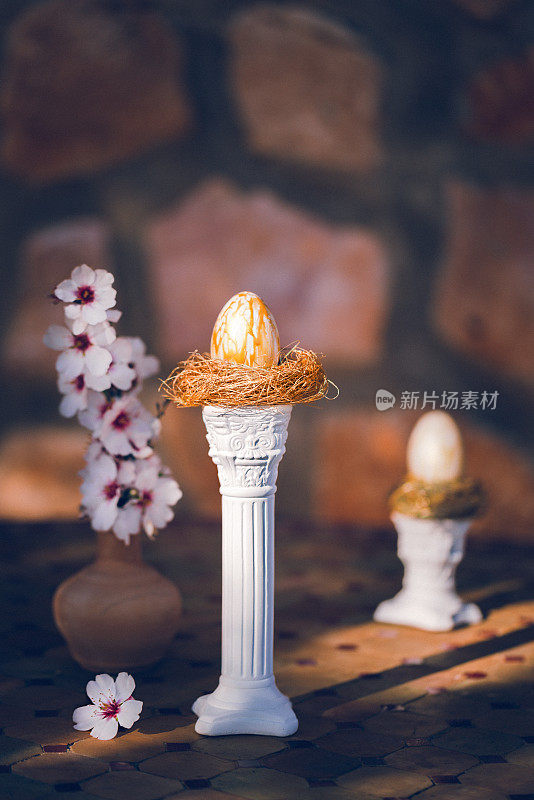在一张古老的乡村桌子上，白色柱子上的金色鸟巢里有两个金色的复活节彩蛋，杏仁花在阳光和阴影的结合中