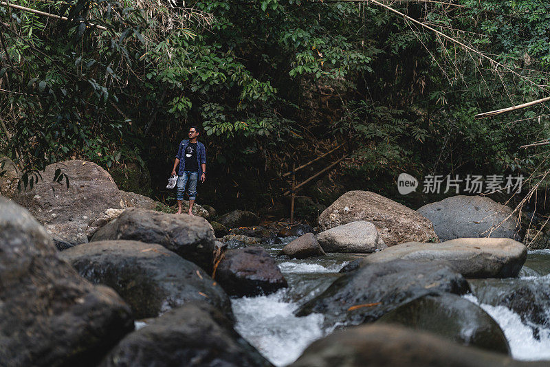 图为，在热带森林中布满岩石的河岸旁，一名亚洲男子正穿着鞋子欣赏着河流的美景