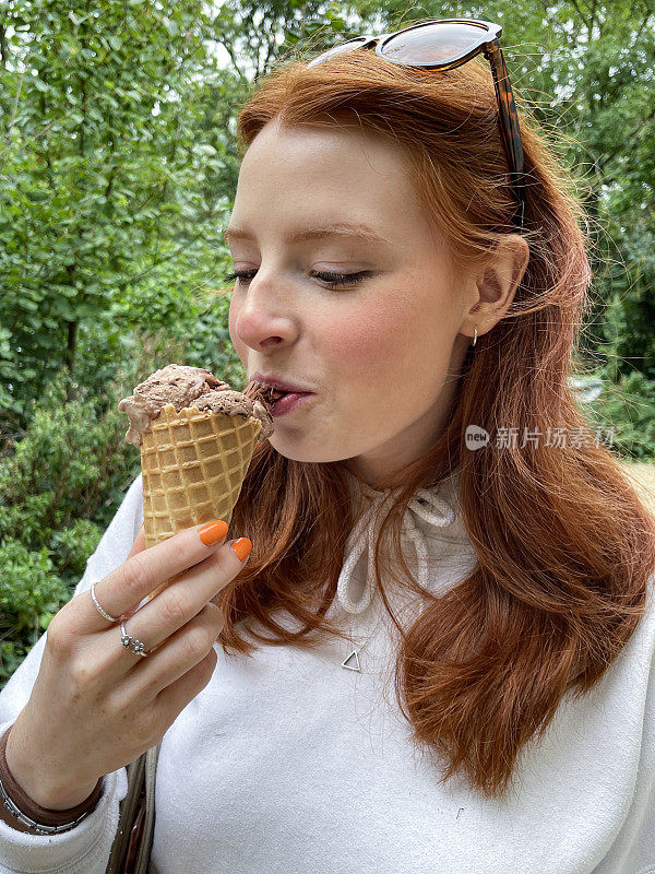 特写照片中，迷人的红发女子头戴墨镜，涂着橙色指甲油，手里拿着一勺巧克力冰淇淋和巧克力片，吃着破碎的巧克力，正在融化的冰淇淋，背景是林地，侧面视图