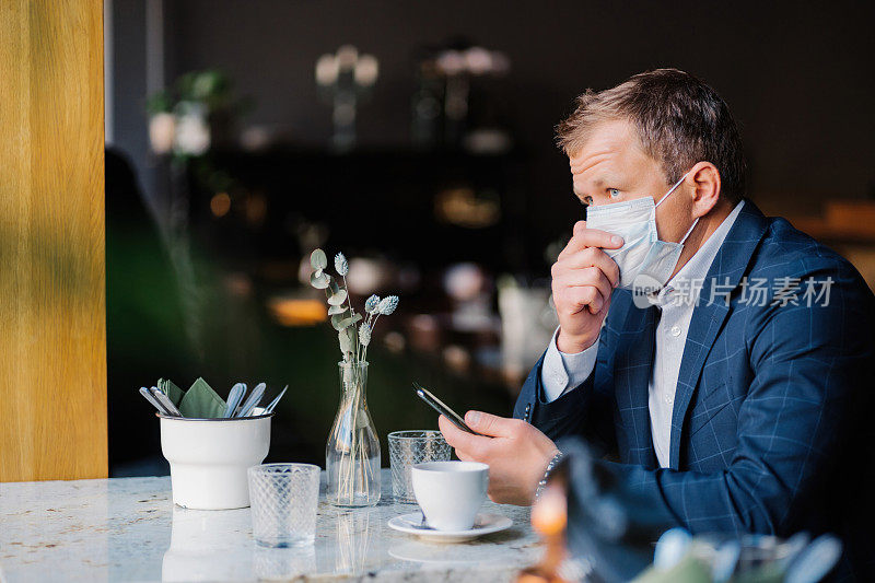 商人在cafÃ©，小心翼翼地摘下口罩喝咖啡，拿着智能手机，新冠疫情期间的新常态反映在他的日常生活中。