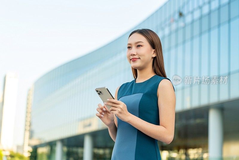 年轻的亚洲女商人在现代化的办公大楼前使用智能手机，带着自信的微笑看向别处