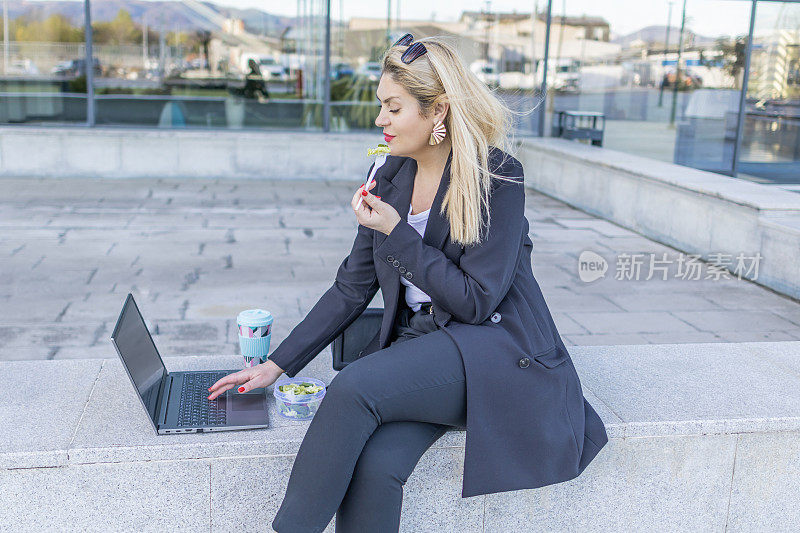 在户外的午餐休息时间，一位微笑的女商人正在吃健康的沙拉。在户外灵活工作。人们在使用科技的同时享受城市的绿色空间。