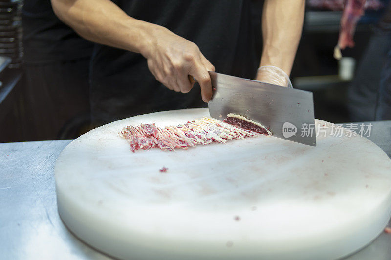 在一家牛肉火锅餐厅的开放式厨房里，厨师在挂着的切碎的生肉后面用刀切牛肉