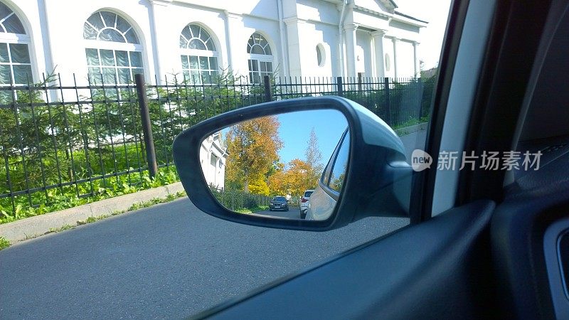 城市街道通过汽车侧后视镜。透过车窗的城市景观。乘坐汽车。橙色和黄色的树在倒影中。公路旅行。