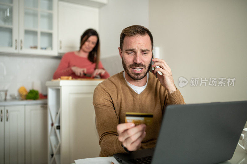 一个年轻的自由职业者正坐在桌子旁打电话。他手里拿着一张信用卡。他面前是他的笔记本电脑。他可爱的妻子跟在他后面，在厨房里切菜。