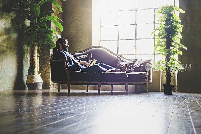 一位留着大胡子的西班牙裔商人躺在办公室后面的沙发上，用平板电脑放松——商业、人与技术概念