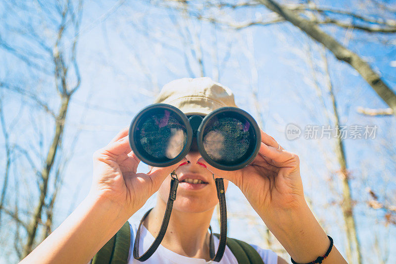 美丽的黑发女孩徒步旅行者。她戴着帽子，背着背包，用望远镜看着相机