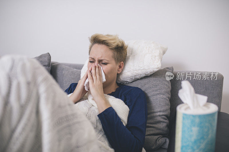 一个生病的年轻女子因感冒坐在家里的沙发上，她盖着一条毯子，擤着鼻子