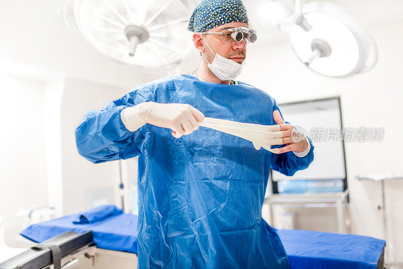 在医院手术室里做美容手术的男性外科医生。外科医生戴口罩和外科医生放大镜，同时摘掉橡胶手套