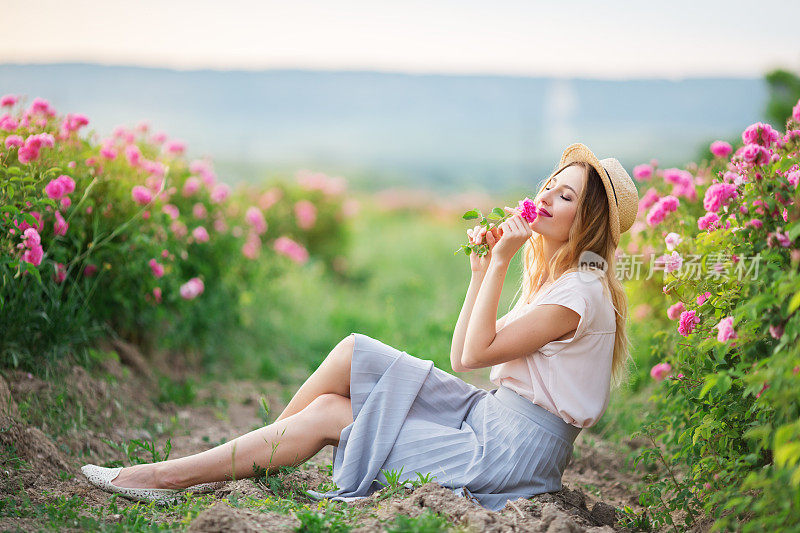美丽的年轻女孩戴着草帽坐在花园里，周围有粉红色的玫瑰