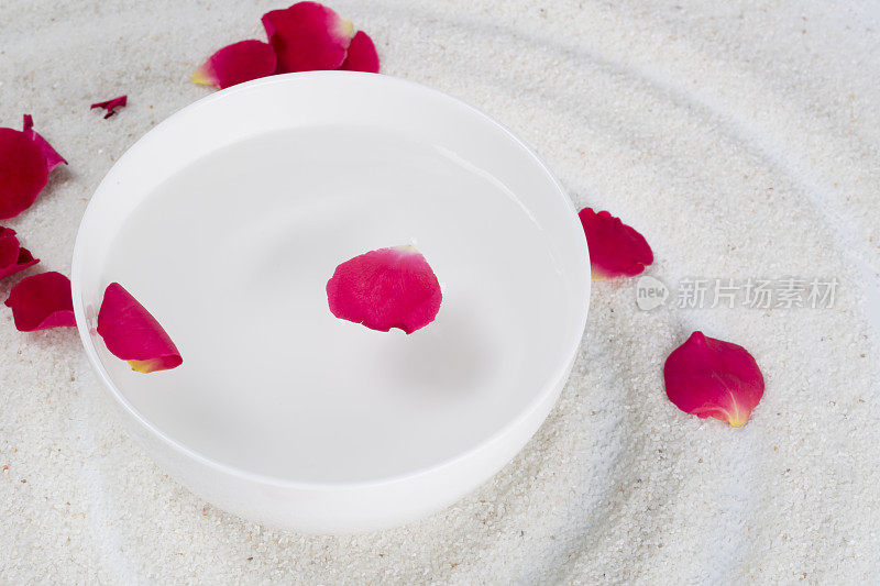 白色的碗里放着玫瑰花瓣，还有白色的沙子。水疗中心。