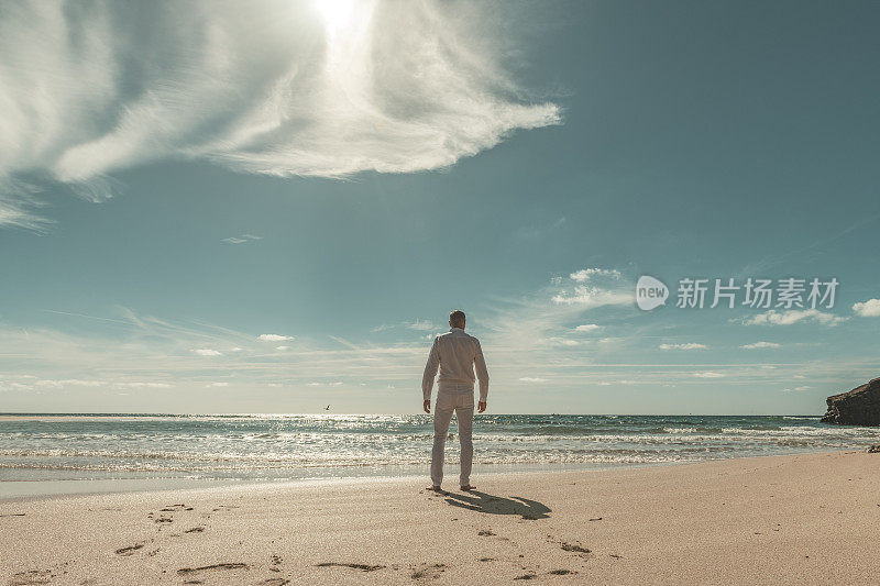穿着一身白衣服的人在阳光明媚空旷的海滩上走向水边。