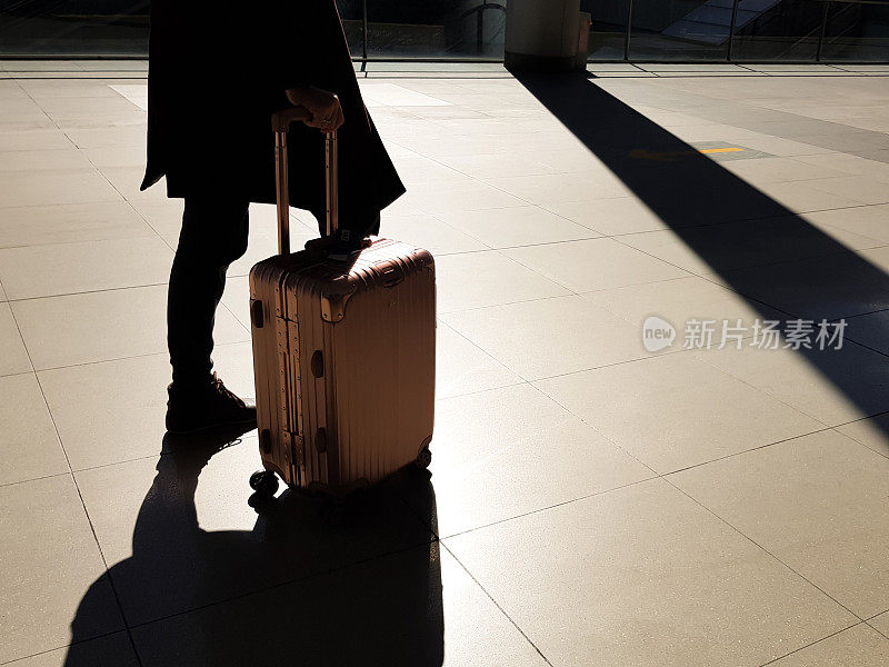 剪影拍摄的女人穿着长礼服冬衣滚动行李在机场航站楼起飞前与早晨的金色阳光时刻。旅游度假或商务工作概念。