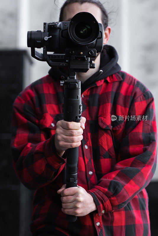年轻的专业摄像师手持专业摄像机，安装在三轴框架稳定器上。专业设备有助于制作高质量的视频而不摇晃。一名身穿红衬衫的摄影师正在拍摄录像。