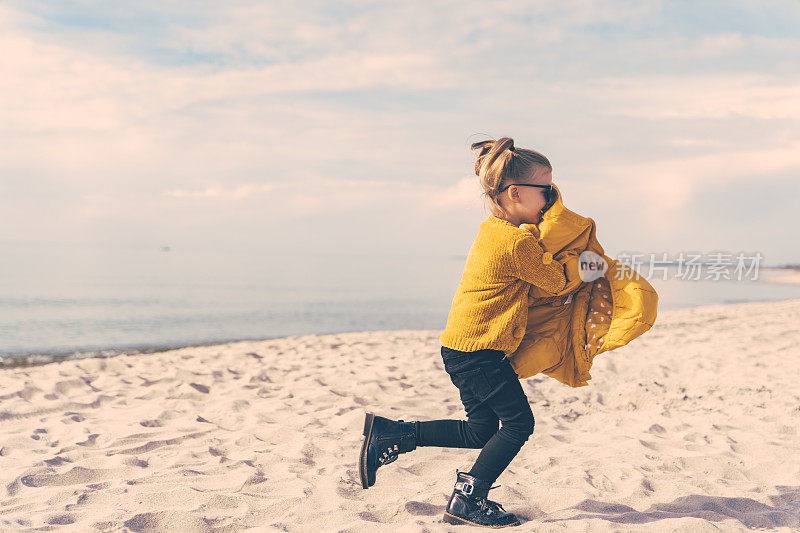 一个梳着有趣马尾辫的5岁小女孩在沙滩上跑步时脱下了外套。