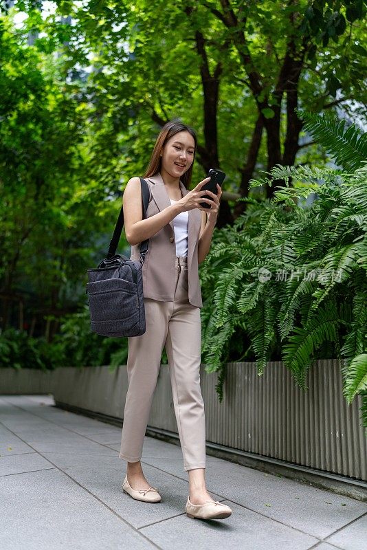 移动中的商业。自然、平衡和互联的移动性。一位年轻的亚洲女商人背着笔记本电脑包，用智能手机上下班，她在中央商务区上班，背景是郁郁葱葱的绿树和花园