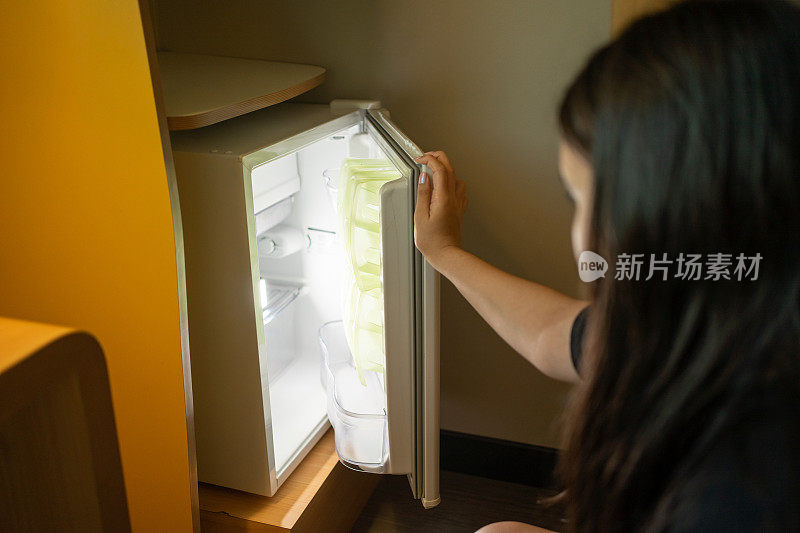 一个女孩打开旅馆里的小冰箱，冰箱里亮着白光。