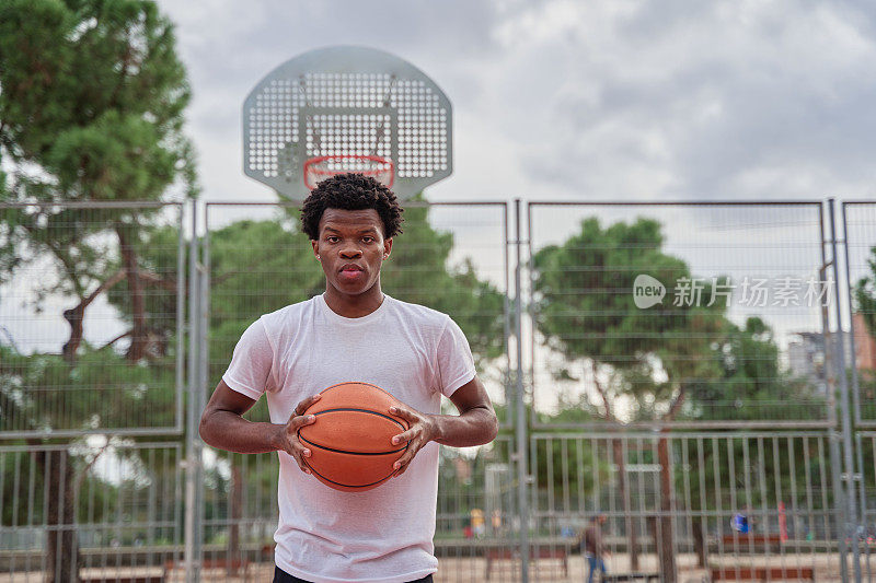 年轻英俊的非裔美国男子篮球运动员双手持球，微笑着看着镜头。有篮球场的城市背景。男篮球运动员的肖像。