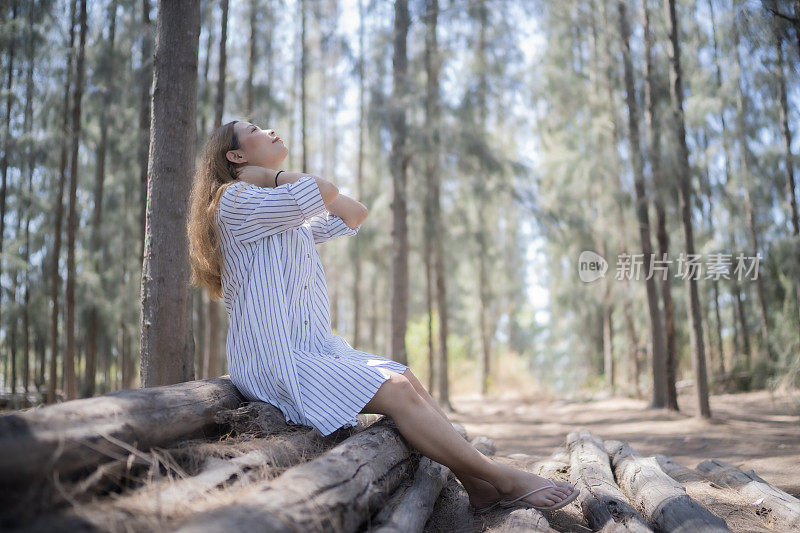 年轻的亚洲女子坐在松林的原木上，仰望，抚摸着自己的脖子，充满了清新，感受自然，风，太阳，意识到自然的重要。