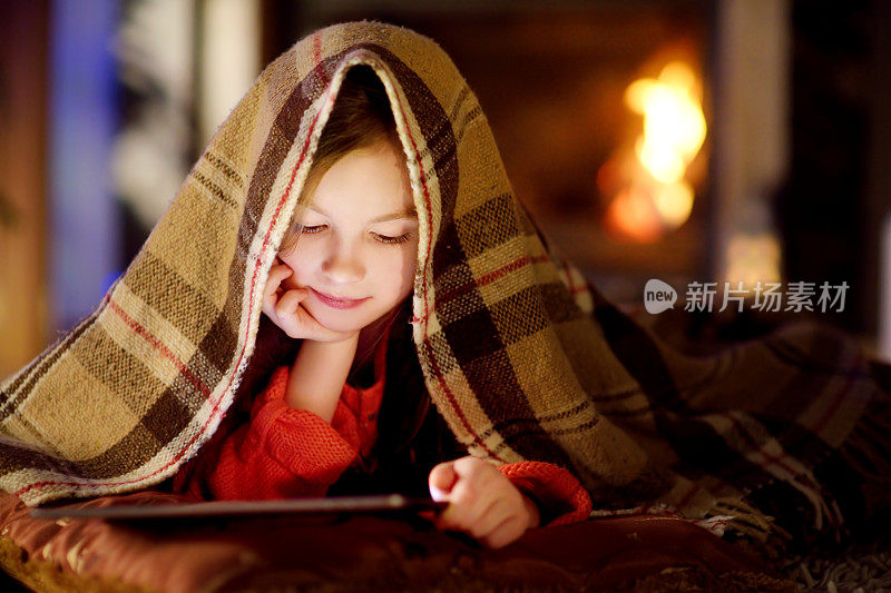 可爱的小女孩在圣诞节晚上壁炉旁使用平板电脑