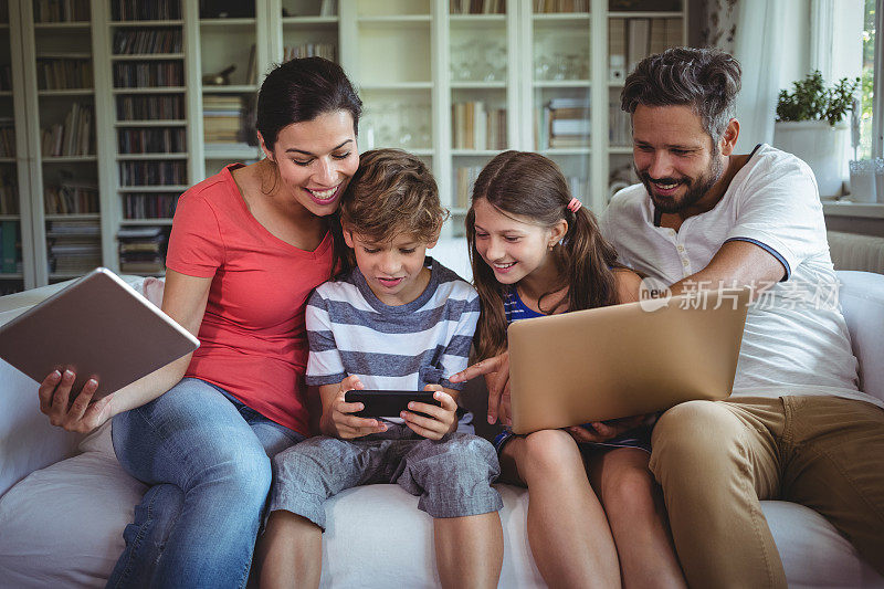 幸福的家庭坐在沙发上，使用笔记本电脑，手机和数字平板电脑
