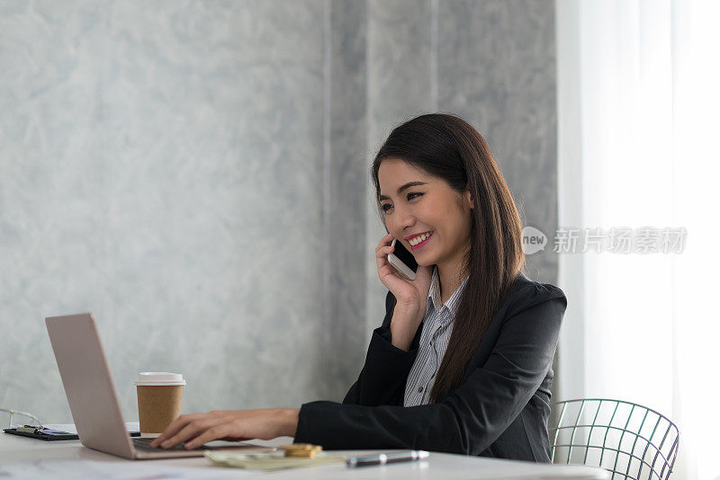 美丽的亚洲年轻女商人在笔记本电脑上工作，而被微笑电话在她的办公桌与明亮的窗户在背景