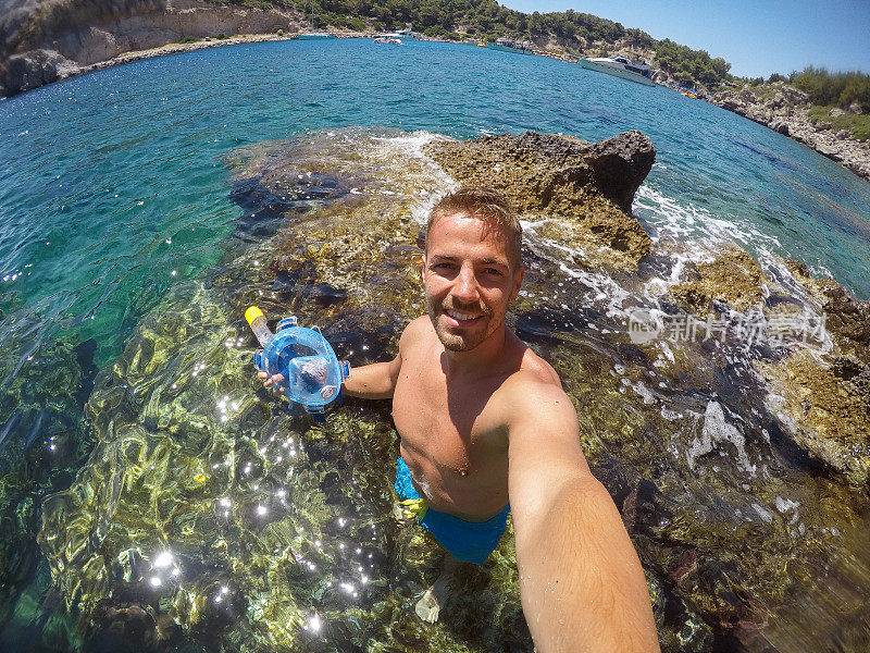 英俊的年轻快乐的游客男子享受夏天，并与一个移动的自拍，而站在海上的石头上，拿着潜水面罩。
