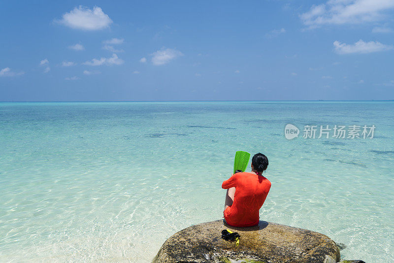 后视图的年轻妇女坐在岩石上，而在海滩上的蓝色的天空和蓝绿色的水，马尔代夫