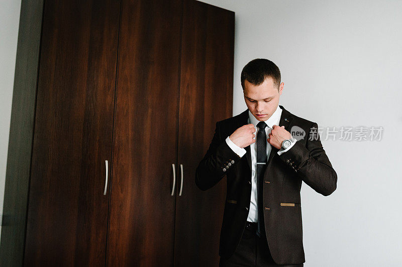 那人正在家里整理夹克。新郎穿着衬衫，打着领带站在房间里。关闭了。帅气的男人模型在正式的西装。服装的概念。新郎在婚礼的早晨。