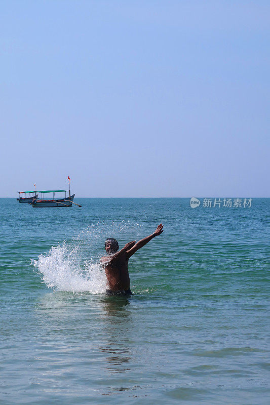 印度一名男子在海滩度假，站在齐腰深的水中，在印度洋中溅起水花，戴着护目镜，伸开双臂溅起水花，看起来很开心，印度果阿的帕洛伦海滩
