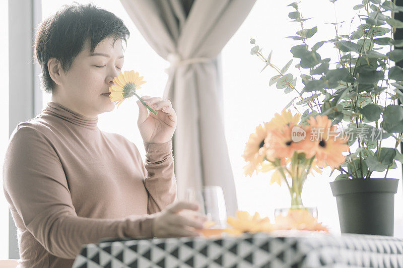 一位亚洲华人中年妇女在她的公寓里享受她的早晨，非洲菊闻着它