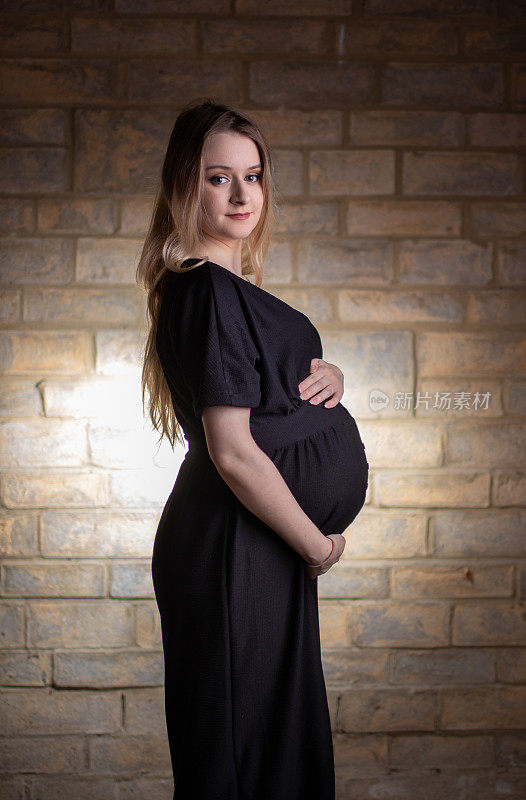 这是一幅以砖墙为背景拍摄的快乐、漂亮、年轻的怀孕妈妈的肖像，她有着金色的长发。
