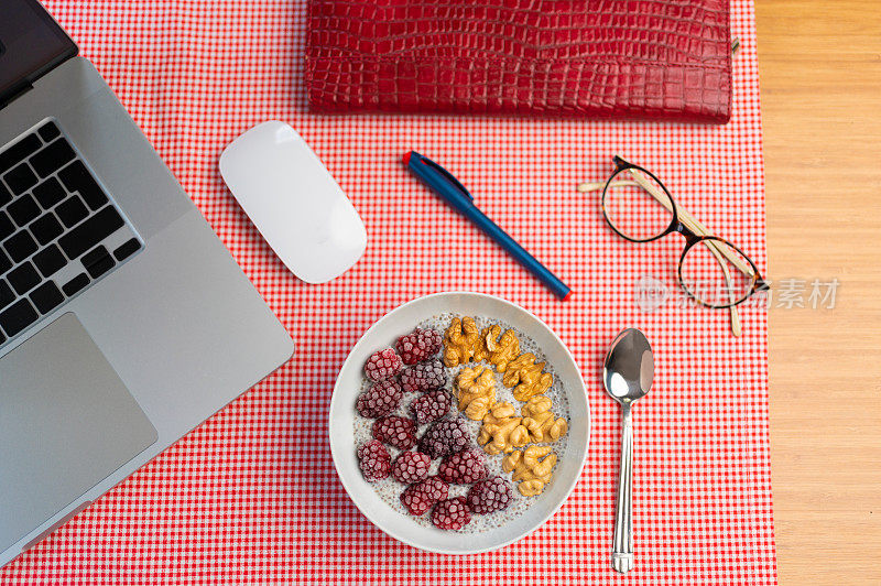 俯视图的健康午餐碗与奇亚，树莓和核桃，打开笔记本电脑，笔，笔记本，眼镜在家庭办公室工作。在家工作的时候，吃点健康的零食休息一下。