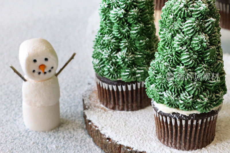 这是一堆自制的圣诞树设计的巧克力纸杯蛋糕，放在木制的蛋糕架上，顶部是覆盖着绿色黄油糖霜的冰淇淋锥，上面撒着糖霜，还有棉花糖雪人