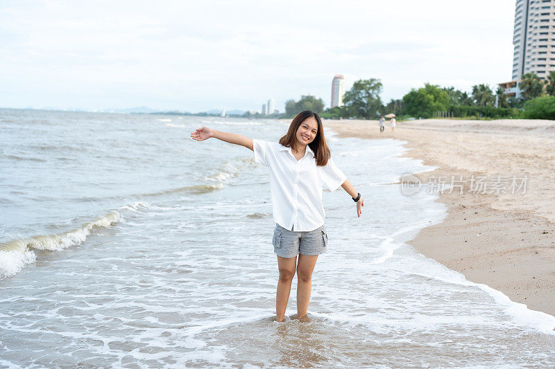 身穿白衬衫、长发的亚洲女性在周末度假时，正带着愉快的表情走在海浪不断拍打的海滩上。日落时的傍晚气氛