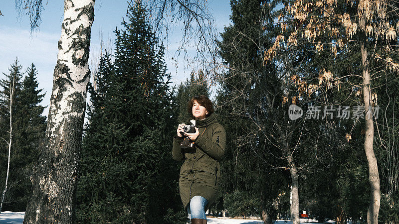 一名女子在阳光明媚的冬日公园散步时，正在使用她的老式胶卷相机